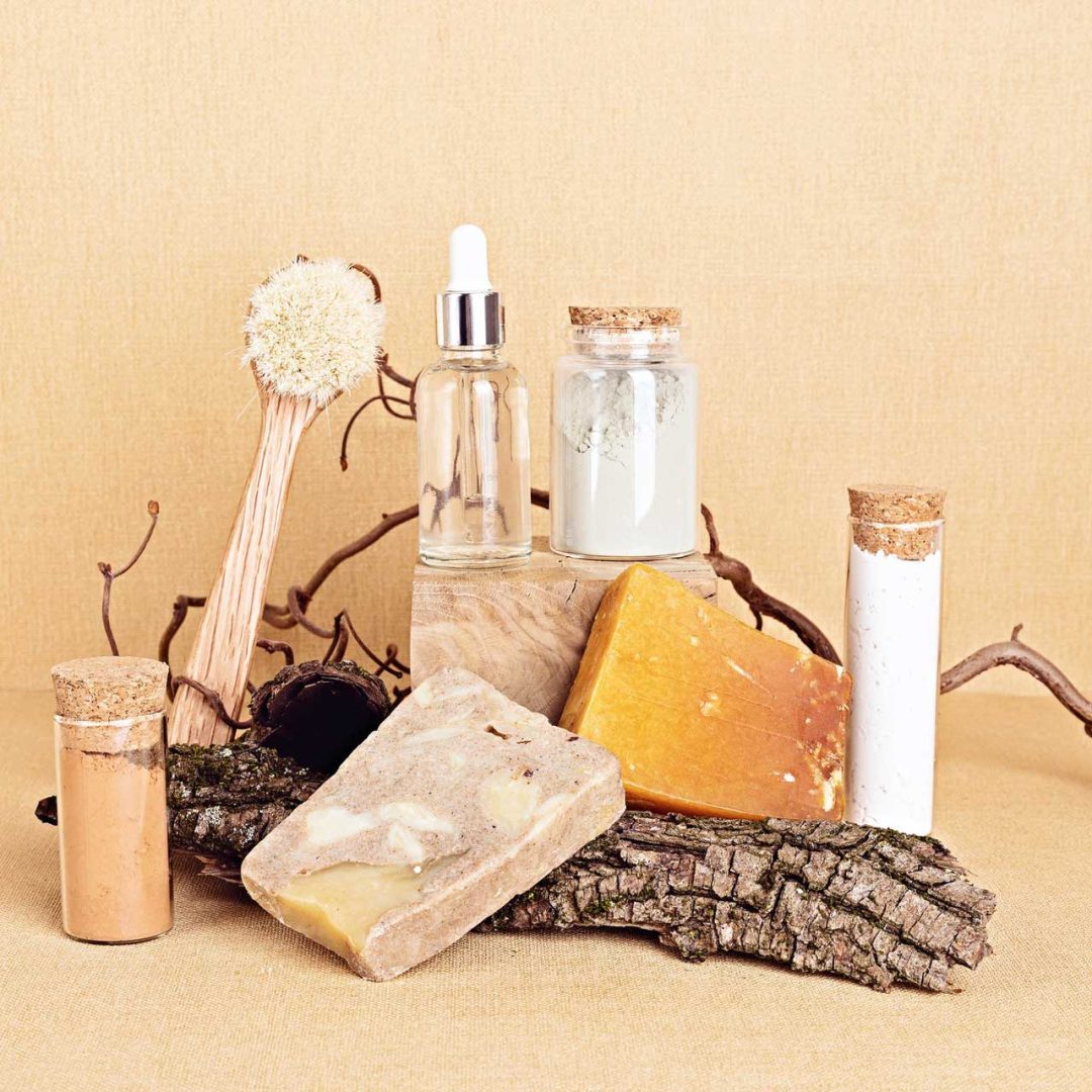 natural-bristle-body-brush-with-serum-soap-and-clay-masks-displayed-with-pieces-of-wood-on-sand-e1655284635779.jpg